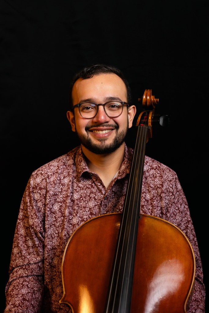 A portrait photo of Franklin Sandoval and his cello. He is smiling and wearing a maroon, pink, and white, densley floral long-sleeve button-up shirt. He has a beard and short brown hair.