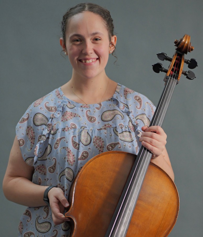 A portrait photo of Grace Raper holding her cello. She is smiling and wearing a light blue paisley patterned dress and has her hair back with pearl earrings and hair ringlets near her ears.