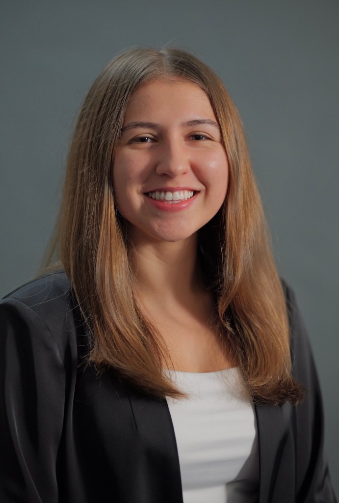A portrait photo of Megan Herrman. She is smiling and wearing a black blaizer and white shirt. Her long auborn hair is down.